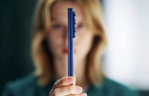 A person holding a blue Samsung Galaxy Z Fold7 smartphone in profile, showcasing its ultra-thin folded design with camera module visible; the background is softly blurred to emphasize the device's sleek form factor